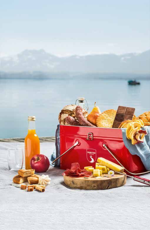 The red Grand Tour Snack-Box, filled with Swiss picnic specialties beside a lake.